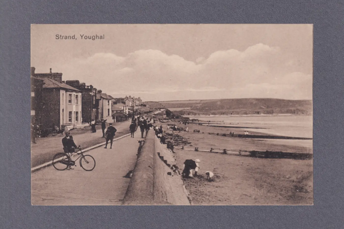 A sepia-toned postcard showing the seafront at Youghal, with people on the beach, promenaders and a man with a bicycle.